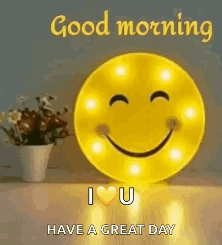 A Smiley Face Is Sitting On A Table Next To A Vase Of Flowers And A Good Morning Message . GIF