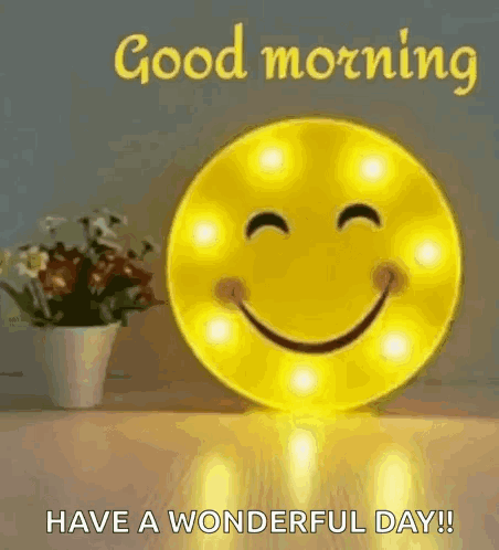 A Smiley Face Is Sitting On A Table Next To A Vase Of Flowers And A Good Morning Message . GIF