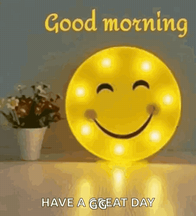 A Smiley Face Is Sitting On A Table Next To A Vase Of Flowers And Says `` Good Morning Have A Great Day '' . GIF
