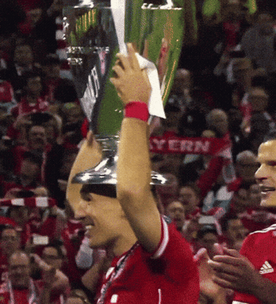 A Soccer Player Holds Up A Trophy In Front Of A Crowd With A Bayern Scarf Behind Him GIF