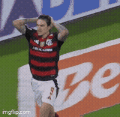 A Soccer Player In A Red And Black Jersey Stands On A Field In Front Of A Sign That Says Be GIF