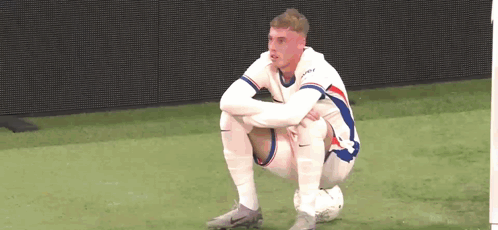 A Soccer Player Is Squatting On The Field With His Legs Crossed And Looking At The Camera . GIF
