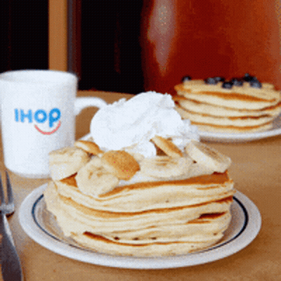 A Stack Of Pancakes With Bananas And Whipped Cream On A Plate Next To An Ihop Mug GIF