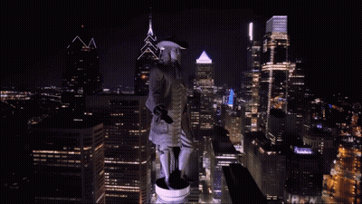 A Statue Of A Man In A Hat Stands On Top Of A City Skyline At Night GIF