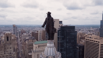 A Statue Of A Man Stands On Top Of A Building In A City With A Chase Bank Building In The Background GIF