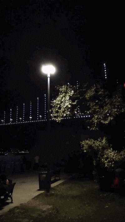 A Street Light Is Lit Up At Night With A Few People Sitting On Benches In The Background GIF