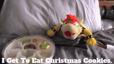 A Stuffed Animal Laying On A Bed Next To A Tray Of Christmas Cookies GIF