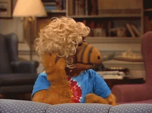 A Stuffed Animal Wearing A Wig And A Blue Shirt Sits On A Couch GIF