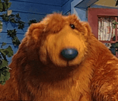 A Stuffed Bear With A Blue Nose Is Standing In Front Of A Blue House GIF