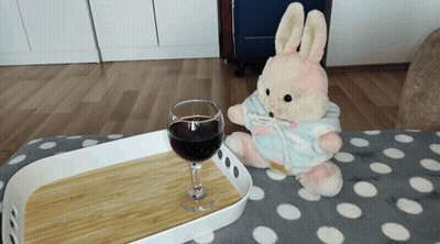 A Stuffed Bunny Is Sitting Next To A Glass Of Wine On A Tray GIF