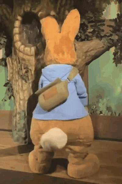 A Stuffed Bunny Is Standing Next To A Tree Wearing A Blue Jacket And A Green Bag . GIF