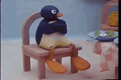 A Stuffed Penguin Is Sitting On A Wooden Chair In Front Of A Table . GIF