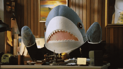 A Stuffed Shark In A Suit Is Sitting At A Desk With Its Mouth Open GIF