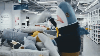 A Stuffed Shark Is Standing In A Store With Pillows GIF