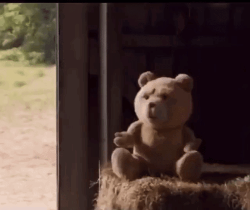 A Teddy Bear Is Sitting On A Bale Of Hay In Front Of A Window . GIF