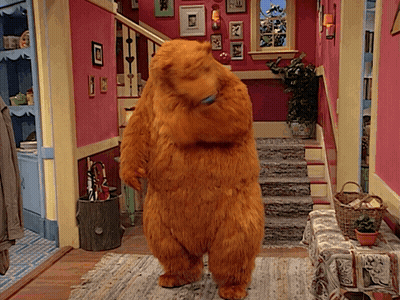 A Teddy Bear Is Standing In A Hallway With Stairs In The Background GIF