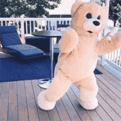 A Teddy Bear Is Standing On A Deck With A Table And Chairs In The Background GIF