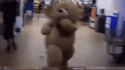A Teddy Bear Is Walking Down A Hallway In A Store With A Person Behind It . GIF