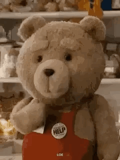 A Teddy Bear Wearing A Red Apron With A Help Button On It Is Standing In Front Of A Refrigerator . GIF
