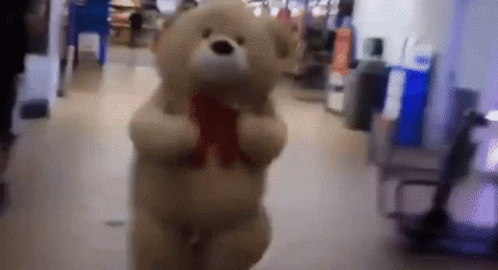 A Teddy Bear With A Red Bow Around Its Neck Is Walking Down A Hallway GIF