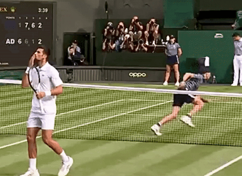 A Tennis Player Is Talking On A Cell Phone While Another Player Is Running Towards The Net . GIF