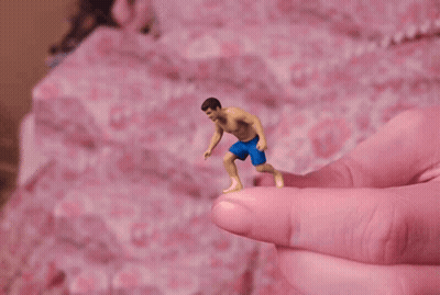 A Tiny Figure Of A Man In Blue Shorts Is Being Held By A Pink Finger GIF
