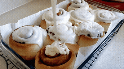 A Tray Of Cinnamon Rolls With White Frosting Being Poured On Them GIF