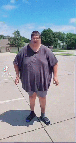 A Very Fat Man Is Standing On A Sidewalk Wearing A Gray Shirt And Sandals . GIF