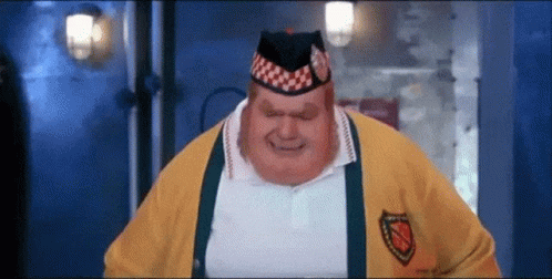 A Very Fat Man Wearing A Yellow Sweater And A Hat Is Standing In A Dark Room . GIF