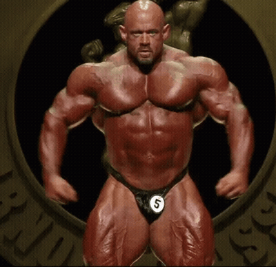 A Very Muscular Man Is Standing In Front Of A Statue Wearing A Number 5 . GIF