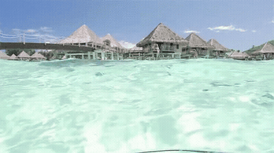 A View Of A Tropical Island From The Water With Thatched Houses In The Background GIF