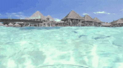 A View Of A Tropical Island From The Water With Thatched Houses In The Background GIF