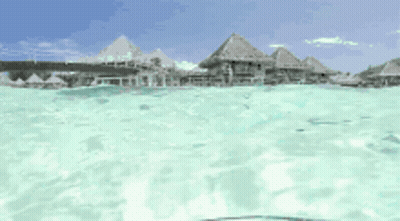 A View Of A Tropical Island From The Water With Thatched Huts In The Background GIF