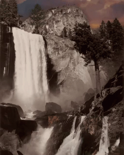 A Waterfall Is Surrounded By Rocks And Trees And A Mountain In The Background GIF