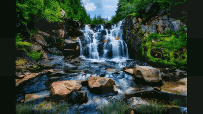 A Waterfall Surrounded By Rocks And Trees With A Person Standing In The Background GIF