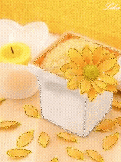 A White Box With A Yellow Flower On It Sits On A Table Next To A Yellow Candle GIF