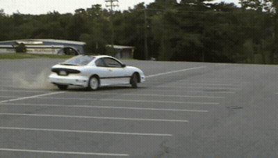 A White Car Is Drifting In A Parking Lot With A Building In The Background GIF