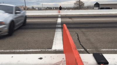 A White Car Is Driving Down A Road With A Red Barrier In The Foreground GIF