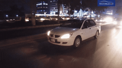 A White Car Is Driving Down A Street At Night With A Pakwheels.com Logo Behind It GIF