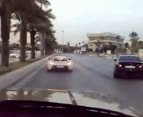 A White Car Is Driving Down A Street Next To A Black Car . GIF