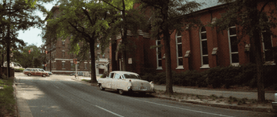 A White Car Is Parked On The Side Of The Road In Front Of A Red Brick Building GIF