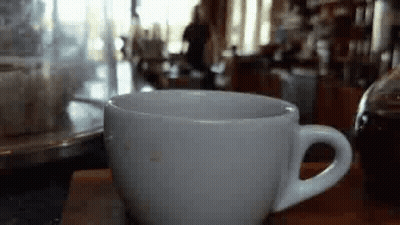 A White Cup Of Coffee Is Sitting On A Wooden Table In A Cafe . GIF