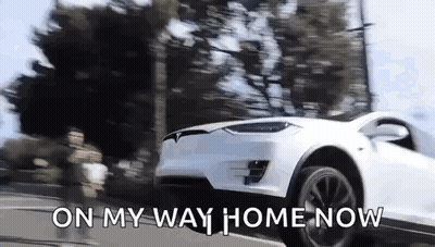 A White Tesla Model X Is Driving Down A Street With The Words `` On My Way Home Now '' Written On The Bottom . GIF