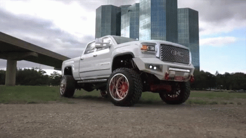A White Truck With Red Wheels Is Parked In A Field In Front Of A Building . GIF