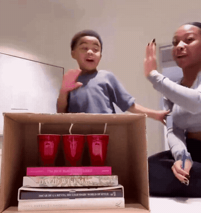 A Woman And A Boy Are Playing With A Cardboard Box Containing A Stack Of Books Including One By Oscar De La Rente GIF