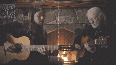 A Woman And An Older Man Are Playing Guitars In Front Of A Fireplace GIF