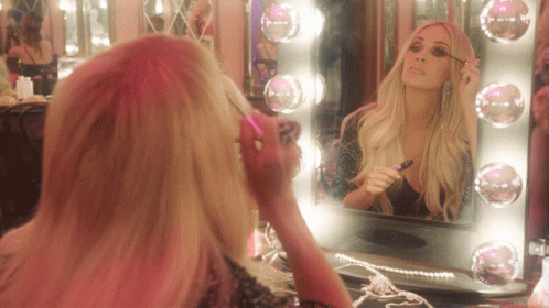 A Woman Applying Mascara In Front Of A Mirror With Pink Lights GIF