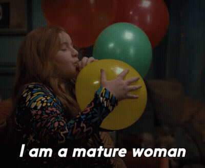A Woman Blowing Up A Yellow Balloon With The Words " I Am A Mature Woman " Below Her GIF