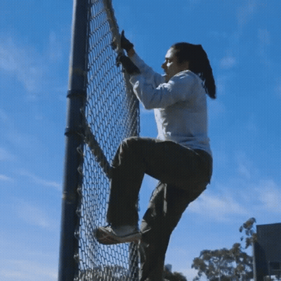 A Woman Climbs A Chain Link Fence Against A Blue Sky GIF