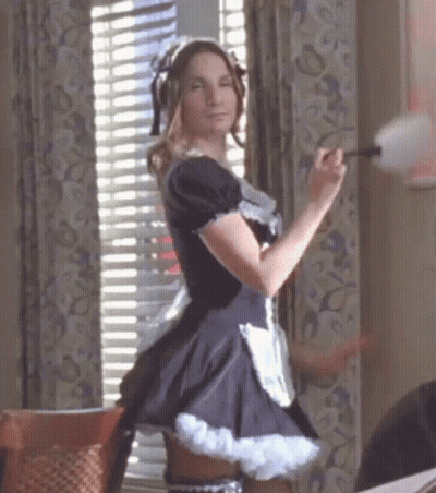A Woman Dressed As A Maid Is Standing In Front Of A Window Holding A Duster . GIF